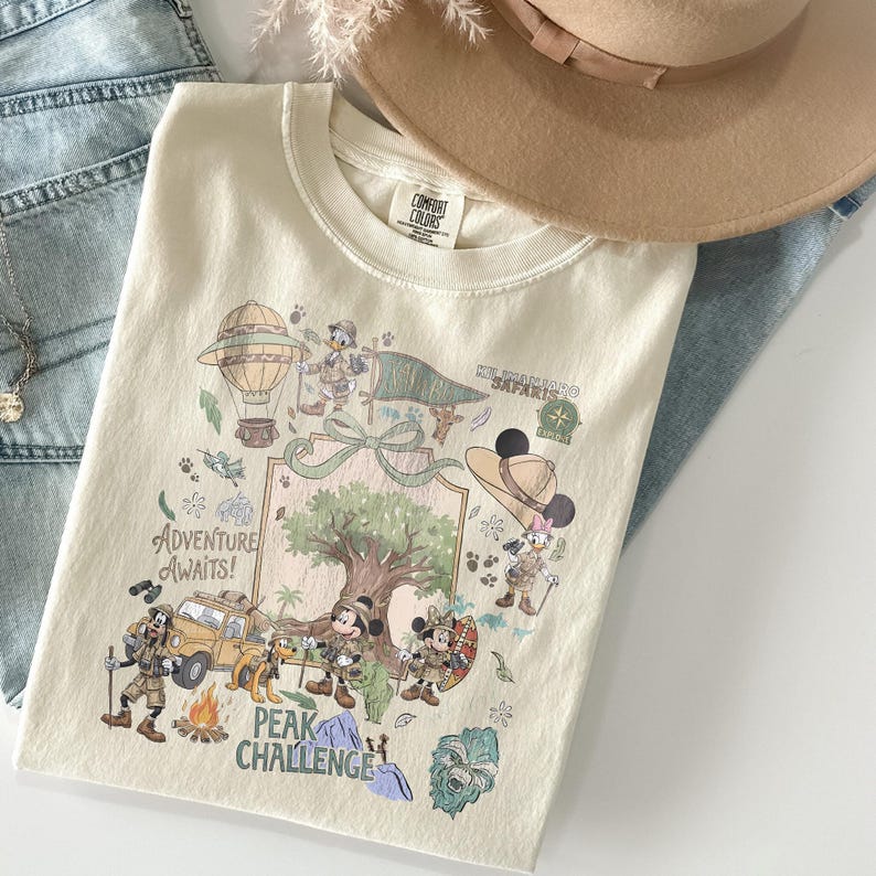 May include: Cream-colored t-shirt featuring a colorful graphic with cartoon characters, a hot air balloon, and the text "Adventure Awaits!" and "Peak Challenge." The shirt is folded on top of blue jeans and a tan hat.