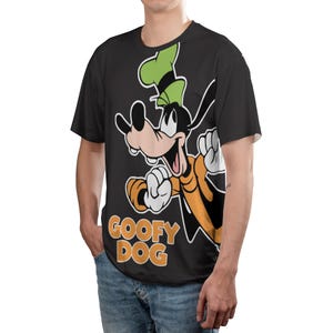 May include: Black t-shirt featuring a cartoon image of Goofy Dog in orange and black, with a green hat. The text "GOOFY DOG" is printed in orange below the character. The shirt is worn by a person.
