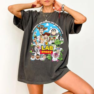May include: Dark gray t-shirt featuring a colorful graphic of Toy Story characters in lab coats, with the text "LAB STORY" and "TO INFINITY AND BEYOND FOR OUR PATIENTS". The design includes Woody, Buzz Lightyear, and other characters.