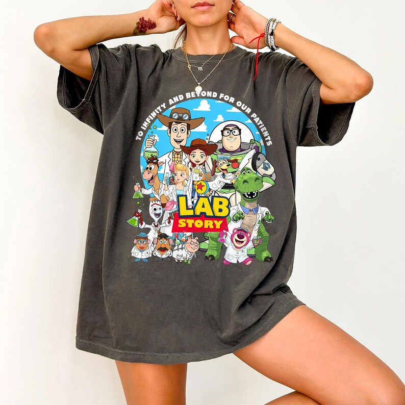 May include: Dark gray t-shirt featuring a colorful graphic of Toy Story characters in lab coats, with the text "LAB STORY" and "TO INFINITY AND BEYOND FOR OUR PATIENTS". The design includes Woody, Buzz Lightyear, and other characters.