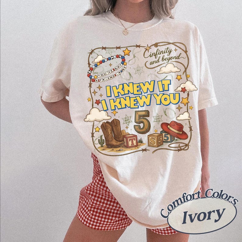 May include: An ivory t-shirt featuring a graphic print with the text "I KNEW IT I KNEW YOU", "Cinfinity and beyond", and "YOU'VE GOT A FRIEND IN ME". The design includes a beaded bracelet, cowboy boots, a red hat, blocks, and stars.
