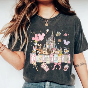 May include: Dark gray t-shirt featuring a colorful Disney World design. The graphic includes Disney characters, a castle, and the word "Disneyworld" in a pastel font. Pink Mickey Mouse balloons and other themed elements are also present.