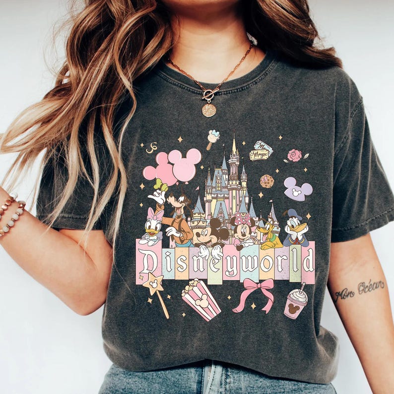 May include: Dark gray t-shirt featuring a colorful Disney World design. The graphic includes Disney characters, a castle, and the word "Disneyworld" in a pastel font. Pink Mickey Mouse balloons and other themed elements are also present.