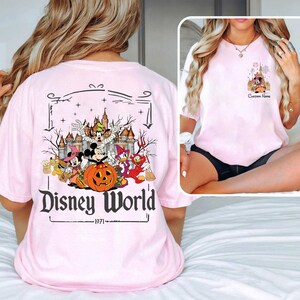 May include: Light pink t-shirt featuring a Disney World Halloween design. The graphic includes Mickey Mouse, Minnie Mouse, Donald Duck, and Goofy, with a pumpkin and castle. The text "Disney World" and "1971" are also visible.