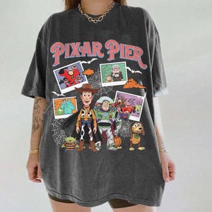 May include: Dark gray t-shirt featuring the words "PIXAR PIER" in pink and red. The shirt has a graphic of various Pixar characters, including Woody, Buzz Lightyear, and others, with a spiderweb design.