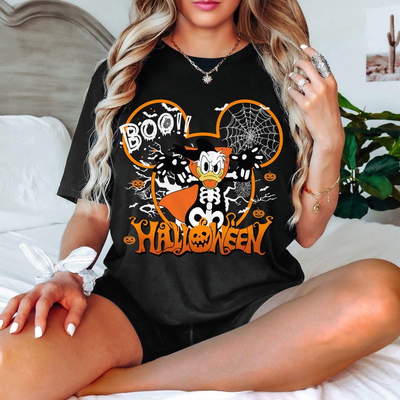 May include: Black t-shirt with a Halloween-themed graphic. The design features Donald Duck dressed as a skeleton with a witch hat, surrounded by a spiderweb, pumpkins, and the words "BOO!!" and "HALLOWEEN" in orange.
