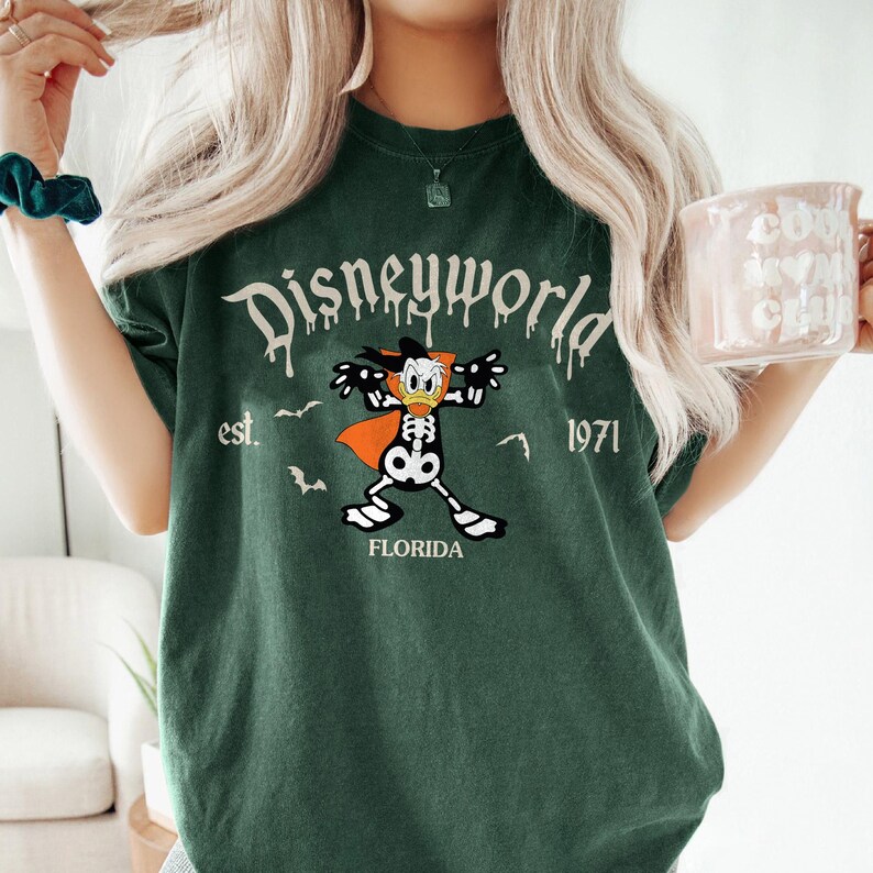 Op de afbeelding: Donkergroen t-shirt met een Disneyworld-afbeelding met een skeletachtig personage met een cape. De tekst "Disneyworld est. 1971 FLORIDA" staat op het shirt. Het shirt heeft een relaxte pasvorm.