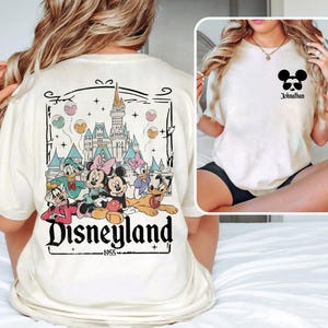 K&ouml;nnte beinhalten: Ein wei&szlig;es T-Shirt mit einer Vintage-Grafik von Mickey Mouse und seinen Freunden vor dem Disneyland-Schloss. Der Text "Disneyland" und "1955" ist unter der Grafik gedruckt. Die R&uuml;ckseite des Shirts zeigt einen schwarzen Mickey-Mouse-Kopf mit dem Namen "Jonathan" darunter.
