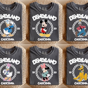 K&ouml;nnte beinhalten: Sechs anthrazitfarbene T-Shirts mit Disney-Figuren. Jedes Shirt zeigt "DISNEYLAND" in Wei&szlig;, mit einer Charakterillustration in einem Kreis, dem Jahr "1955" und "CALIFORNIA". Zu den Charakteren geh&ouml;ren Donald Duck, Mickey Mouse, Goofy, Daisy Duck, Minnie Mouse und Pluto.