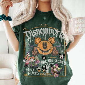 May include: Dark green t-shirt featuring a Halloween-themed graphic. The design includes a smiling pumpkin with Mickey Mouse ears, the words "Disneyworld," "Hocus Pocus," and various Disney characters. The shirt has a vintage, distressed look.