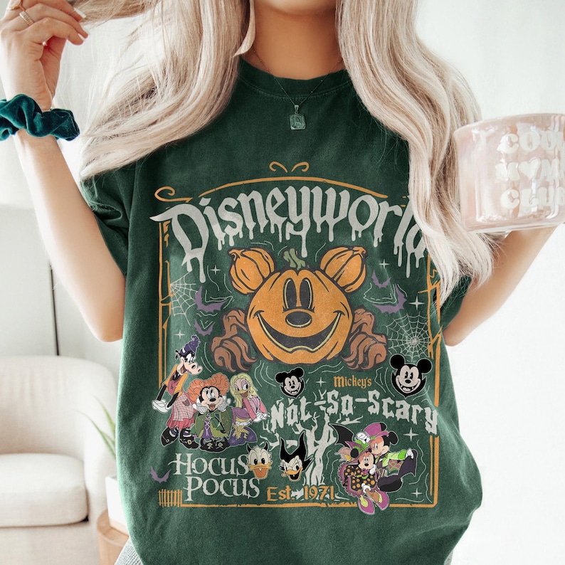 May include: Dark green t-shirt featuring a Halloween-themed graphic. The design includes a smiling pumpkin with Mickey Mouse ears, the words "Disneyworld," "Hocus Pocus," and various Disney characters. The shirt has a vintage, distressed look.