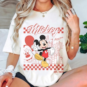 May include: Cream-colored t-shirt featuring a red graphic of Mickey Mouse with the word "Mickey" in a script font. The design includes a castle, fireworks, and the text "est. 1928".