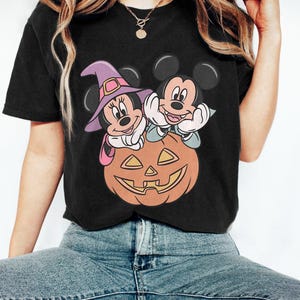 May include: Black t-shirt featuring a Halloween-themed graphic. The design includes Mickey and Minnie Mouse characters, dressed in witch attire, perched atop a carved orange pumpkin. The image has a cartoon style.