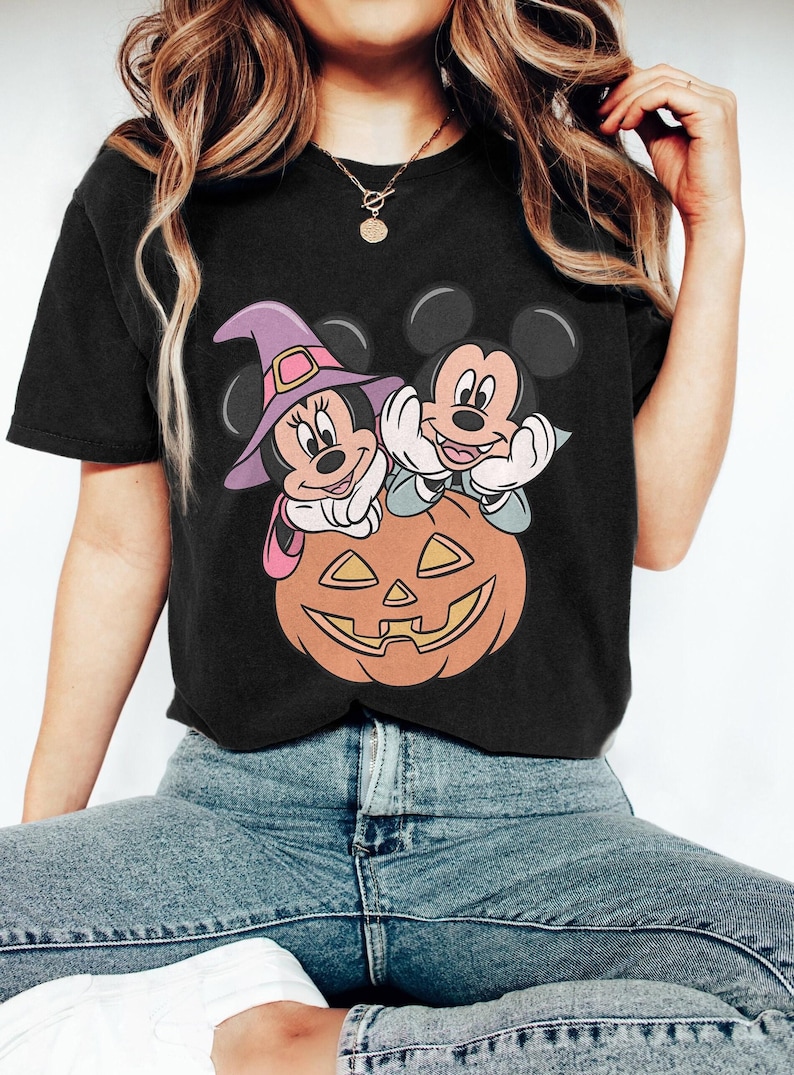 May include: Black t-shirt featuring a Halloween-themed graphic. The design includes Mickey and Minnie Mouse characters, dressed in witch attire, perched atop a carved orange pumpkin. The image has a cartoon style.