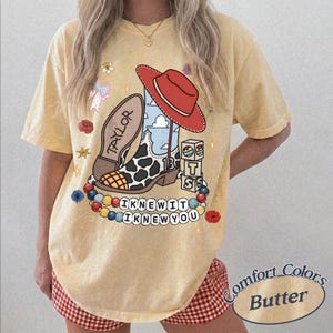 May include: A pale yellow short-sleeve t-shirt featuring a graphic print of a cowboy boot with a cow print pattern, a red cowboy hat, and blocks labelled 13 and TTS. The design includes a string of colourful beads with the text I KNEW IT I KNEW YOU.