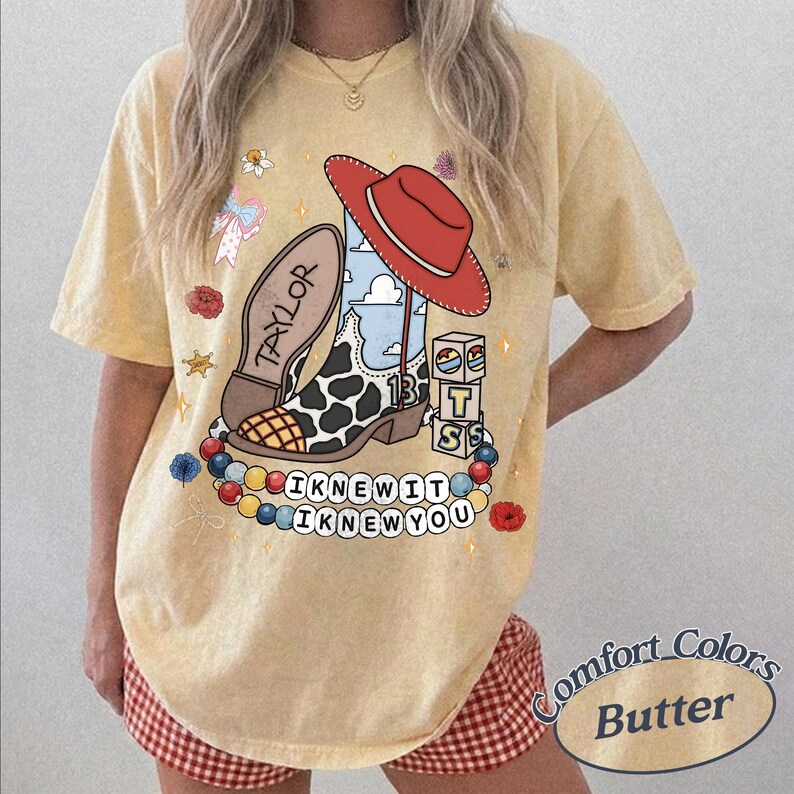 May include: A pale yellow short-sleeve t-shirt featuring a graphic print of a cowboy boot with a cow print pattern, a red cowboy hat, and blocks labelled 13 and TTS. The design includes a string of colourful beads with the text I KNEW IT I KNEW YOU.