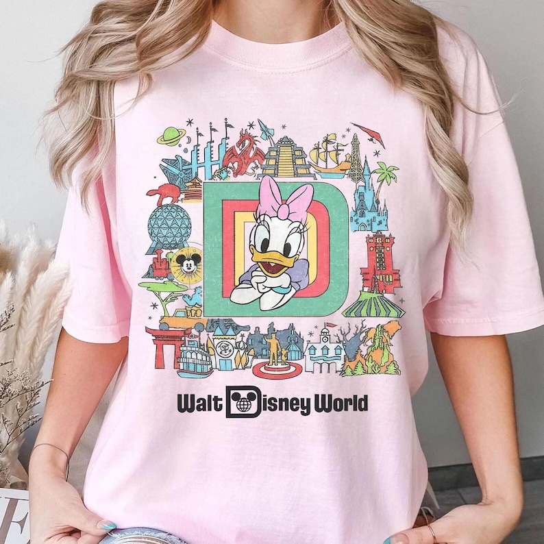 Puede incluir: Camiseta rosa claro con un gr&aacute;fico colorido de Daisy Duck dentro de una gran letra "D", rodeada de im&aacute;genes de Disney World. El texto "Walt Disney World" est&aacute; impreso debajo.