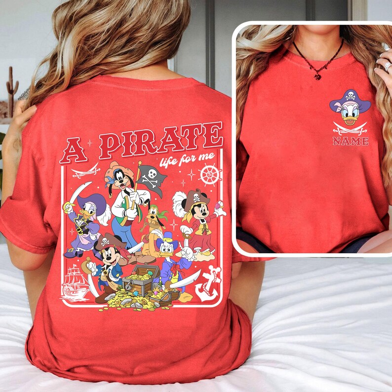 May include: Red t-shirt featuring a pirate-themed graphic with Disney characters. The design includes Mickey Mouse, Donald Duck, Goofy, and others, along with pirate elements like a treasure chest, swords, and a skull and crossbones flag. The text "A Pirate life for me" is also displayed.