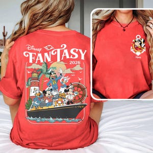 May include: Red t-shirt featuring a Disney Fantasy 2026 design. The back of the shirt displays a graphic of Mickey Mouse, Donald Duck, Goofy, and other characters on a cruise ship. The front has a Mickey Mouse anchor logo.