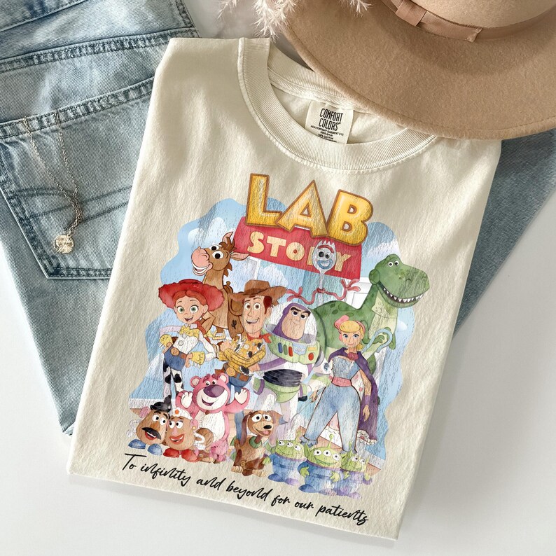 May include: Cream-colored t-shirt featuring a colorful graphic of characters from the movie "Toy Story." The design includes Woody, Buzz Lightyear, and other characters, with the words "LAB STORY" and "To infinity and beyond for our patients" printed on the shirt.