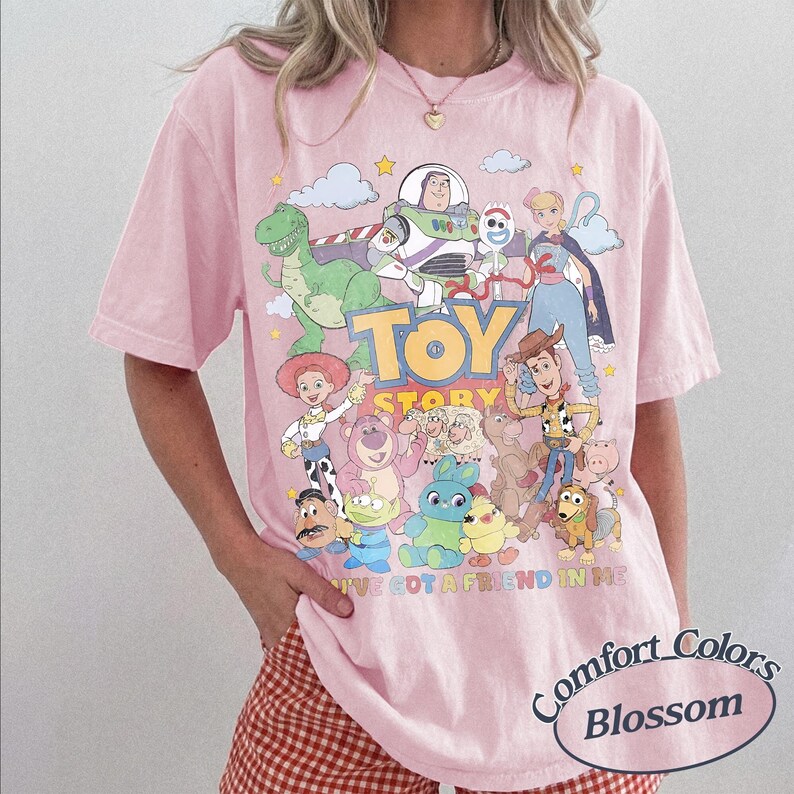May include: A light pink short-sleeve t-shirt featuring a colorful graphic print of Toy Story characters including Buzz Lightyear, Woody, Jessie, and Rex. The text "TOY STORY" and "YOU'VE GOT A FRIEND IN ME" are printed in the center with various clouds and stars.