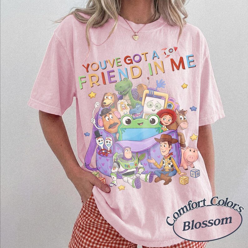 May include: A light pink t-shirt featuring a graphic print of various animated characters from Toy Story emerging from a purple backpack. The text "YOU'VE GOT A FRIEND IN ME" is printed in colorful letters above the characters, with a small rocket icon.