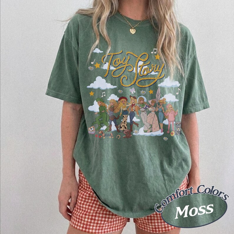 May include: A moss green short-sleeve t-shirt featuring a graphic print of Toy Story characters. The design includes the text Toy Story in yellow rope-style lettering, white clouds, musical notes, and stars above a group of illustrated characters.