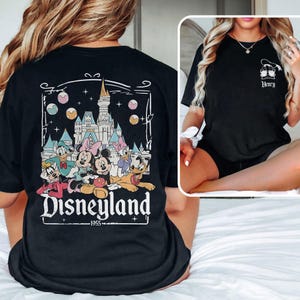 K&ouml;nnte beinhalten: Schwarzes T-Shirt mit einem Aufdruck von Mickey Mouse und seinen Freunden vor dem Disneyland-Schloss. Der Text "Disneyland - 1955" ist unter dem Aufdruck gedruckt.