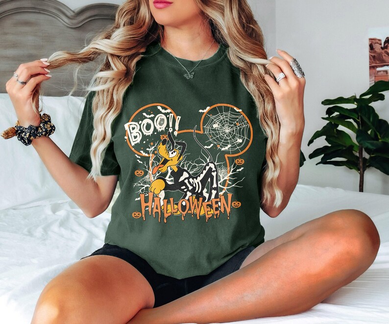 Pode incluir: T-shirt verde escuro com um design de Halloween. O design apresenta um c&atilde;o de desenho animado com uma fantasia de esqueleto, uma teia de aranha e as palavras "BOO!!" e "HALLOWEEN" em laranja. A t-shirt &eacute; de estilo casual.