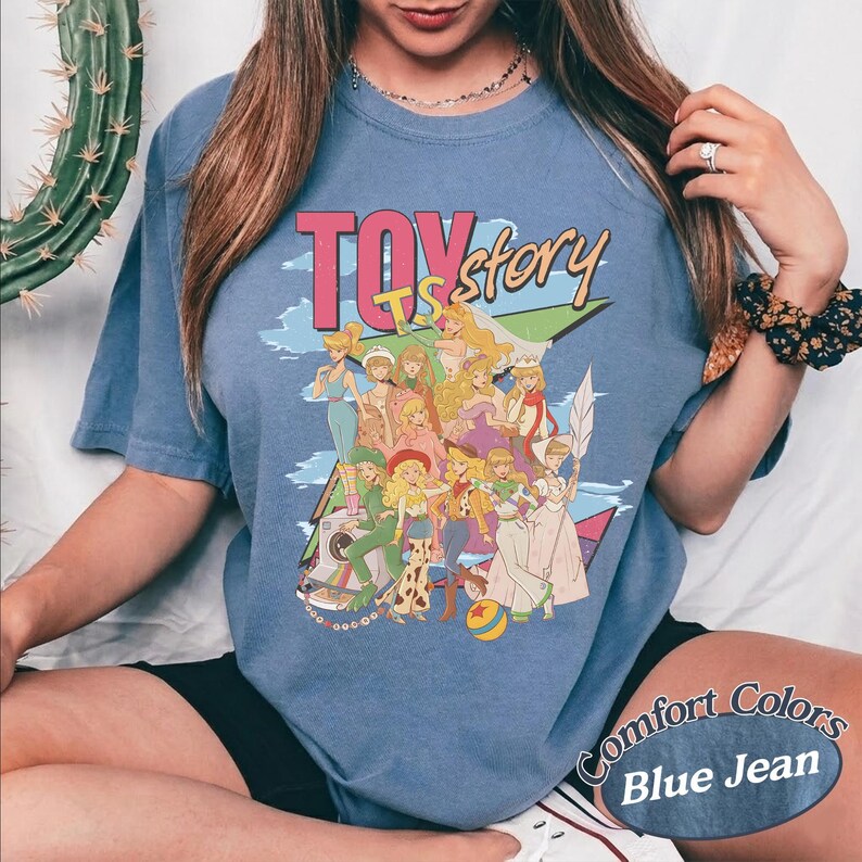 May include: A blue jean colored t-shirt featuring a graphic print with the text TOY story. The design displays a group of illustrated characters in various outfits, including a cowboy hat, a space suit, and a ball with a red star pattern.