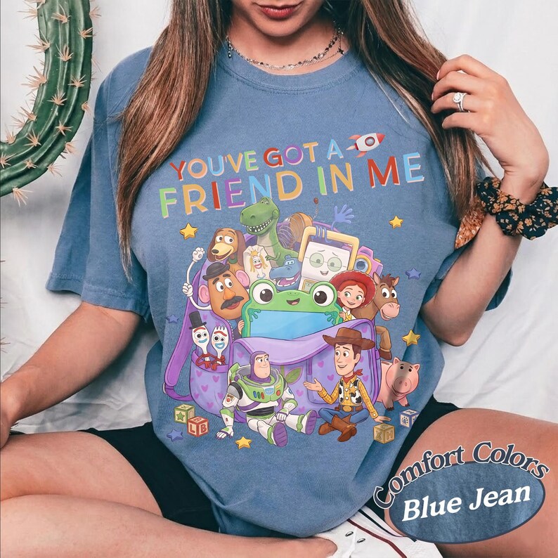 May include: A blue short-sleeve t-shirt featuring a graphic print of various animated characters from the Toy Story franchise emerging from a purple backpack. The text "YOU'VE GOT A FRIEND IN ME" is printed in colorful letters above the characters.