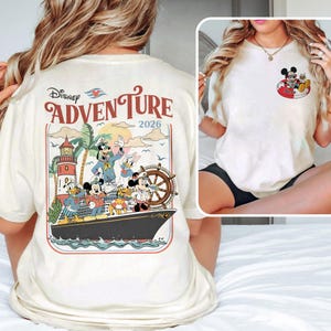 May include: Cream-coloured t-shirt with a Disney Adventure graphic. The design features Mickey Mouse, Donald Duck, Goofy, and Daisy Duck on a cruise ship, with a lighthouse and palm trees. The text "Disney Adventure 2026" is also visible.