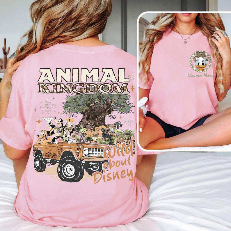 May include: Pink t-shirt with "ANIMAL KINGDOM" text and a graphic of Mickey Mouse, Minnie Mouse, and friends in a safari vehicle. The shirt also features the text "Wild about Disney".
