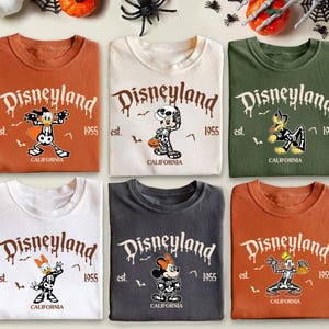 K&ouml;nnte beinhalten: Sechs gefaltete T-Shirts mit Disneyland-Grafiken und Halloween-Charakterdesigns. Die Shirts sind in verschiedenen Farben erh&auml;ltlich, darunter Orange, Wei&szlig; und Gr&uuml;n. Jedes Shirt hat das Disneyland-Logo, das Jahr 1955 und das Wort "California" aufgedruckt.