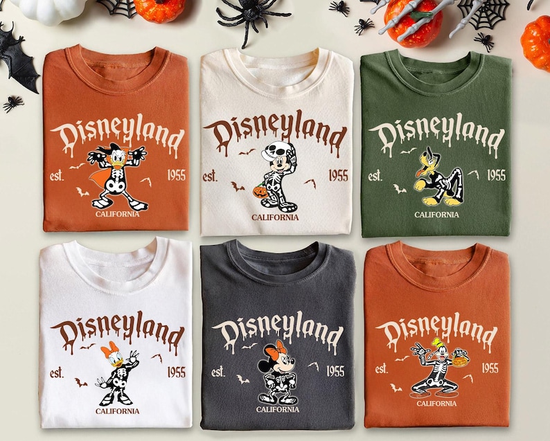 K&ouml;nnte beinhalten: Sechs gefaltete T-Shirts mit Disneyland-Grafiken und Halloween-Charakterdesigns. Die Shirts sind in verschiedenen Farben erh&auml;ltlich, darunter Orange, Wei&szlig; und Gr&uuml;n. Jedes Shirt hat das Disneyland-Logo, das Jahr 1955 und das Wort "California" aufgedruckt.