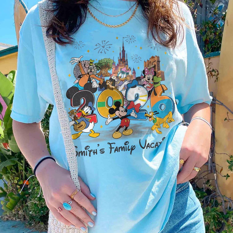 Może przedstawiać: Jasnoniebieska koszulka z kolorową grafiką postaci Disneya, w tym Myszki Miki, Kaczora Donalda i Myszki Minnie, z napisem "2023 Smith's Family Vaca". Koszulka ma kr&oacute;tkie rękawy i okrągły dekolt.