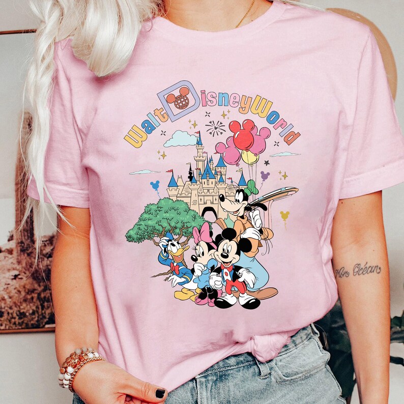 May include: Pink t-shirt with a graphic print of Mickey Mouse, Minnie Mouse, Donald Duck, and Goofy in front of Cinderella's Castle. The text "Walt Disney World" is printed in a rainbow arc above the characters.