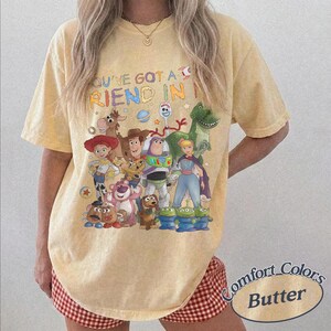 May include: A pale yellow short-sleeve t-shirt featuring a graphic print of various animated characters from the Toy Story franchise. The text "YOU'VE GOT A FRIEND IN ME" is printed in colorful letters above the characters. The shirt has a crew neckline.