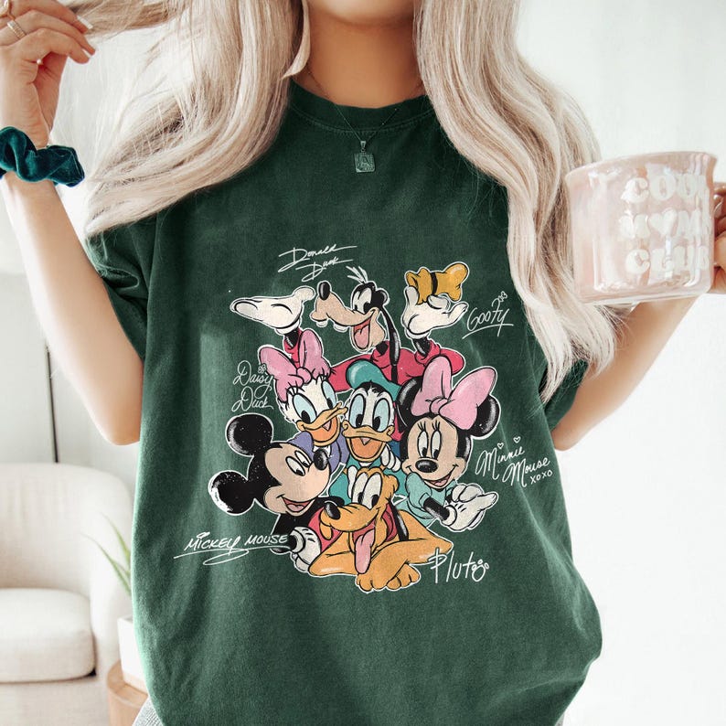 May include: Dark green t-shirt featuring a graphic of Disney characters including Mickey Mouse, Minnie Mouse, Donald Duck, and Goofy. The characters' names are written above or below them in a handwritten style. The shirt is a relaxed fit.