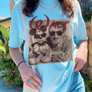 May include: Light blue t-shirt featuring a vintage-style graphic of Mickey Mouse and Walt Disney. The graphic is in black and white with the word "Walt" in red script. The shirt is a casual style.