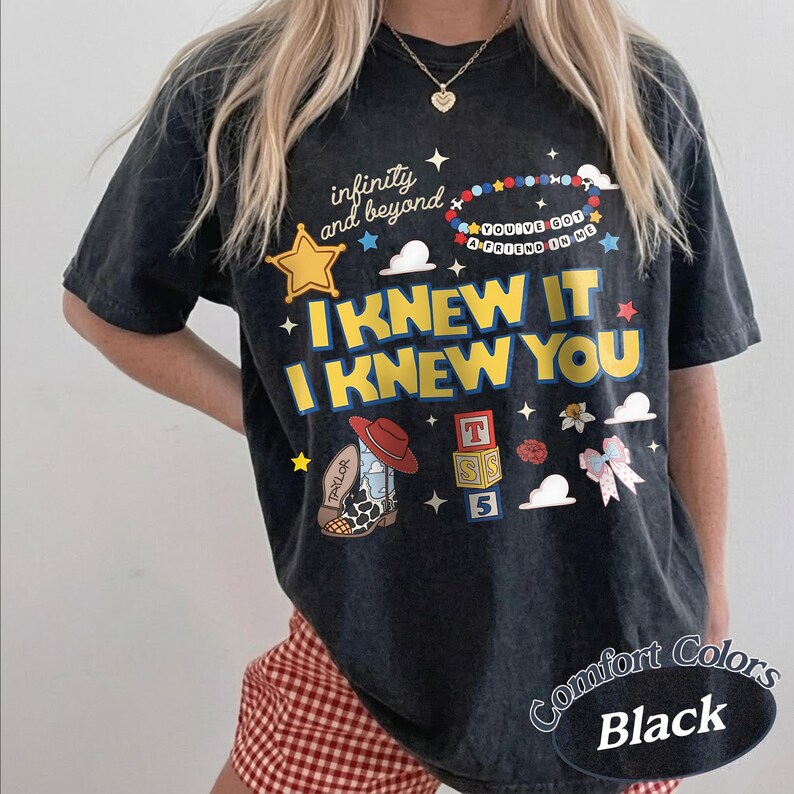 May include: A black short-sleeve t-shirt featuring a graphic print with the text "infinity and beyond", "YOU'VE GOT A FRIEND IN ME", "I KNEW IT", and "I KNEW YOU". The design includes a yellow star, a beaded bracelet, a cowboy boot, blocks, and a bow.
