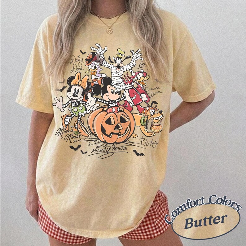 May include: A pale yellow short-sleeve t-shirt featuring a Halloween-themed graphic of Mickey Mouse, Minnie Mouse, Donald Duck, Daisy Duck, Goofy, and Pluto in costumes. The design includes a large orange pumpkin and text labels for each character.