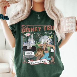Pu&ograve; includere: T-shirt verde scuro con una grafica colorata di Disney Trip 2026. Il design include un castello, Spaceship Earth e Pippo. La maglietta ha anche uno striscione con la scritta "Custom Name" e una testa di Topolino con la scritta "Hollywood Studios".