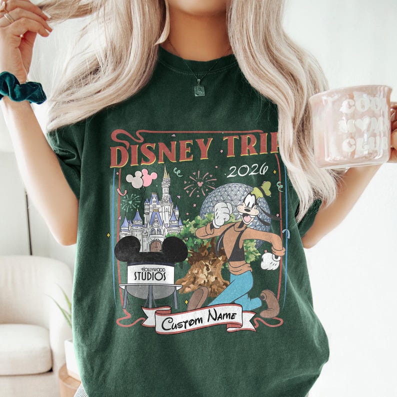 Pu&ograve; includere: T-shirt verde scuro con una grafica colorata di Disney Trip 2026. Il design include un castello, Spaceship Earth e Pippo. La maglietta ha anche uno striscione con la scritta "Custom Name" e una testa di Topolino con la scritta "Hollywood Studios".