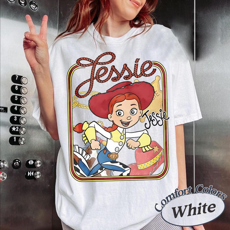 May include: A white t-shirt featuring a graphic print of the character Jessie from Toy Story. The design includes a red cowboy hat, a yellow and white shirt, blue trousers, and a rope border. The name Jessie is written in a rope-style font above.