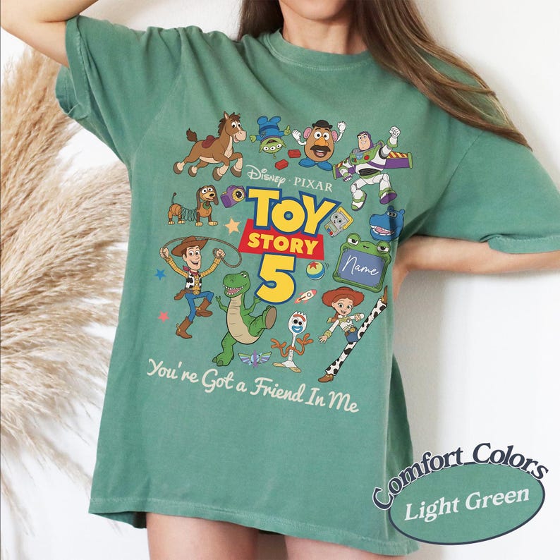 May include: A light green t-shirt featuring a colorful graphic of Toy Story characters including Woody, Buzz Lightyear, Rex, and Jessie. The design includes the text Toy Story 5 and You've Got a Friend In Me, with a blank space for a name.