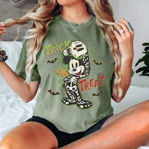 May include: An olive green t-shirt with a Halloween Mickey Mouse design. The graphic shows Mickey in a skeleton costume holding a skull mask, with the words "Trick or Treat" and bat silhouettes.