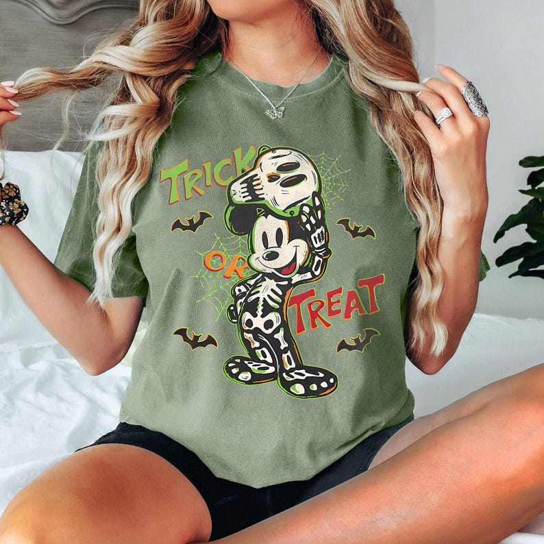 May include: An olive green t-shirt with a Halloween Mickey Mouse design. The graphic shows Mickey in a skeleton costume holding a skull mask, with the words "Trick or Treat" and bat silhouettes.