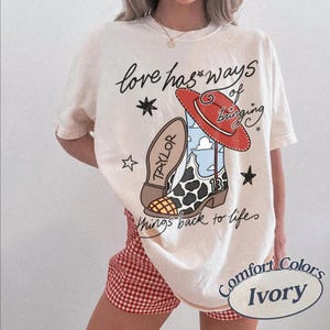 May include: An ivory t-shirt featuring a graphic of a cow-print cowboy boot with a red hat, blue sky pattern, and the text "Love has ways of bringing things back to life" along with the name "TAYLOR" and the number "13" printed on the boot.