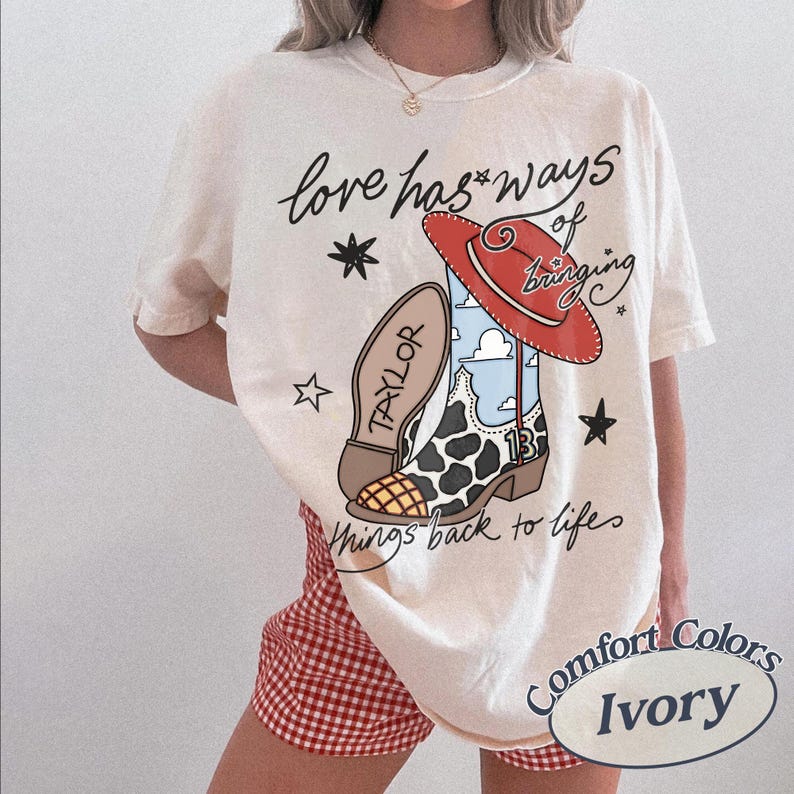 May include: An ivory t-shirt featuring a graphic of a cow-print cowboy boot with a red hat, blue sky pattern, and the text "Love has ways of bringing things back to life" along with the name "TAYLOR" and the number "13" printed on the boot.